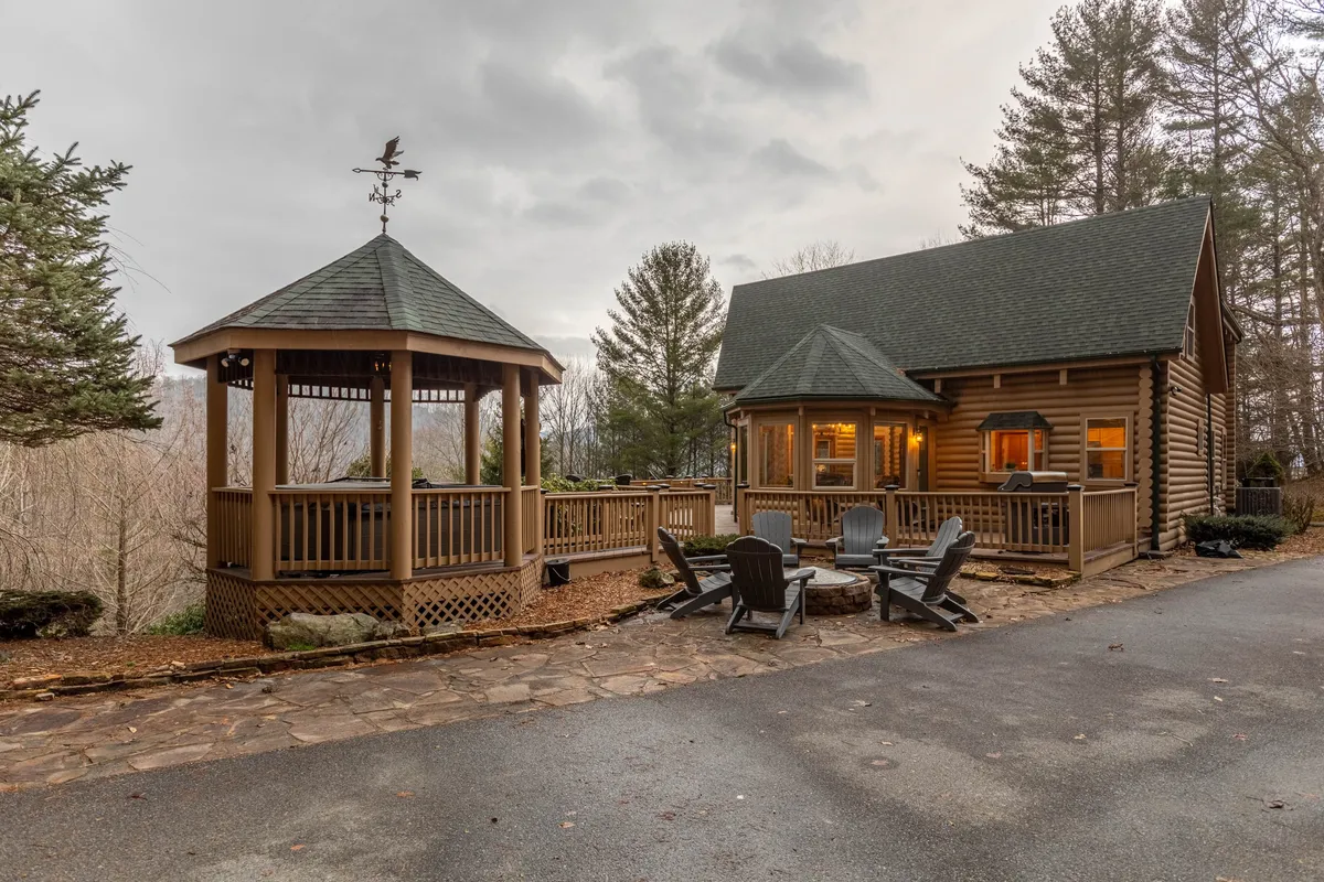 This image showcases the exterior of Whispering Willow, featuring the log cabin, a welcoming gazebo, and a fire pit surrounded by trees. The scene highlights inviting outdoor spaces perfect for family gatherings, story sharing, and relaxing in nature.