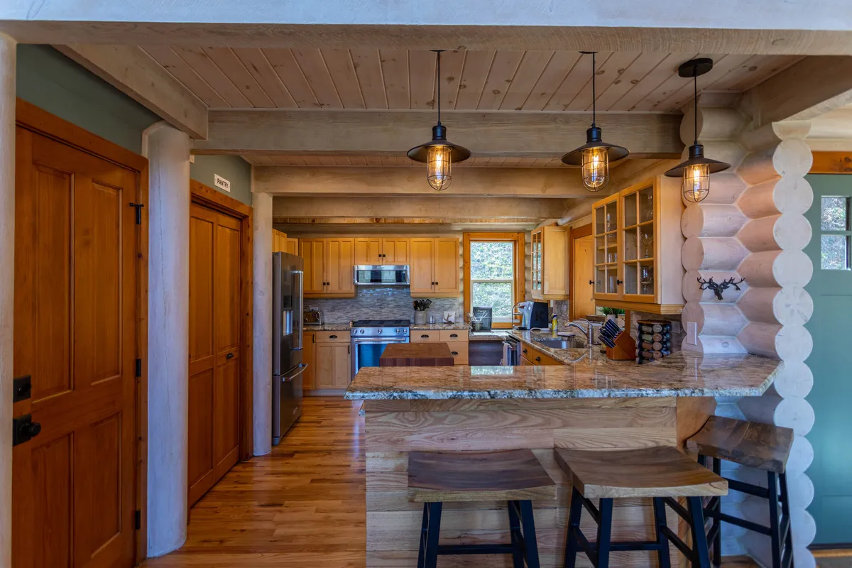 A warm, inviting kitchen showcasing a spacious granite countertop with wooden bar stools, open shelving filled with glassware, and premium stainless steel appliances. The kitchen is well-lit and designed for both cooking and gathering, blending modern convenience with rustic charm.