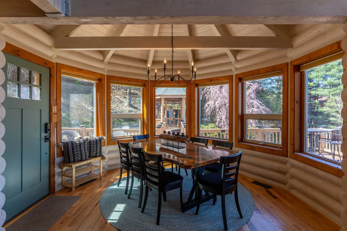 A focus on the dining area with a wooden table for eight, a rustic chandelier overhead, and large windows providing natural light and scenic views. The log walls add character and warmth, making it an inviting spot for shared meals or celebrations.