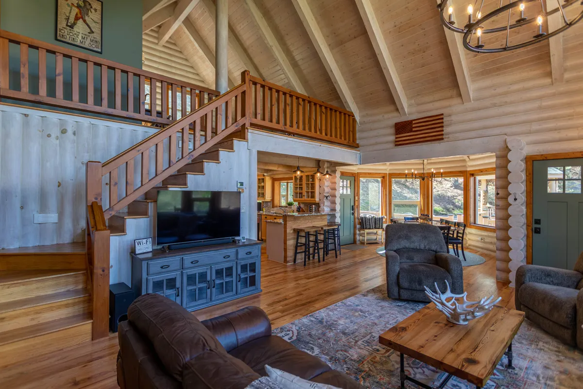 A broad view of the living and dining area reveals a cozy seating arrangement, large windows pouring in sunlight, a visible kitchen area with bar seating, and a rustic staircase leading upstairs. The atmosphere is warm and inviting, ideal for both relaxation and entertaining.