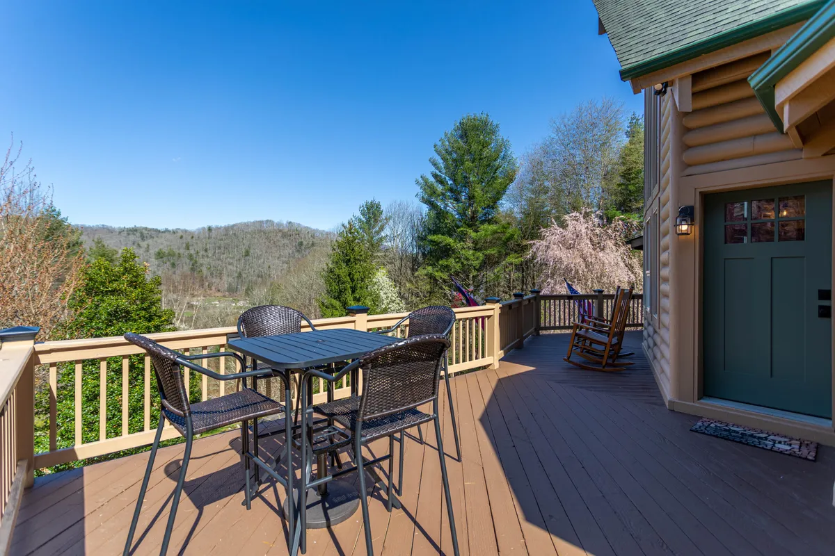 A scenic covered deck with comfortable outdoor table and chairs, wooden railing, and panoramic views of the surrounding mountains. The setting invites peaceful mornings, fresh air, and tranquil moments overlooking nature.