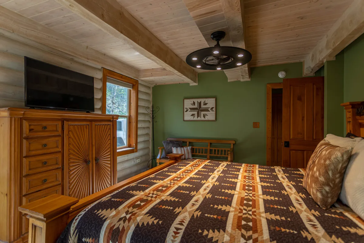 A cozy bedroom with a bed dressed in a patterned blanket, rustic wooden furniture, a Smart TV, and a decorative art piece above the headboard. Natural light streams in from a window, creating a warm and inviting atmosphere.