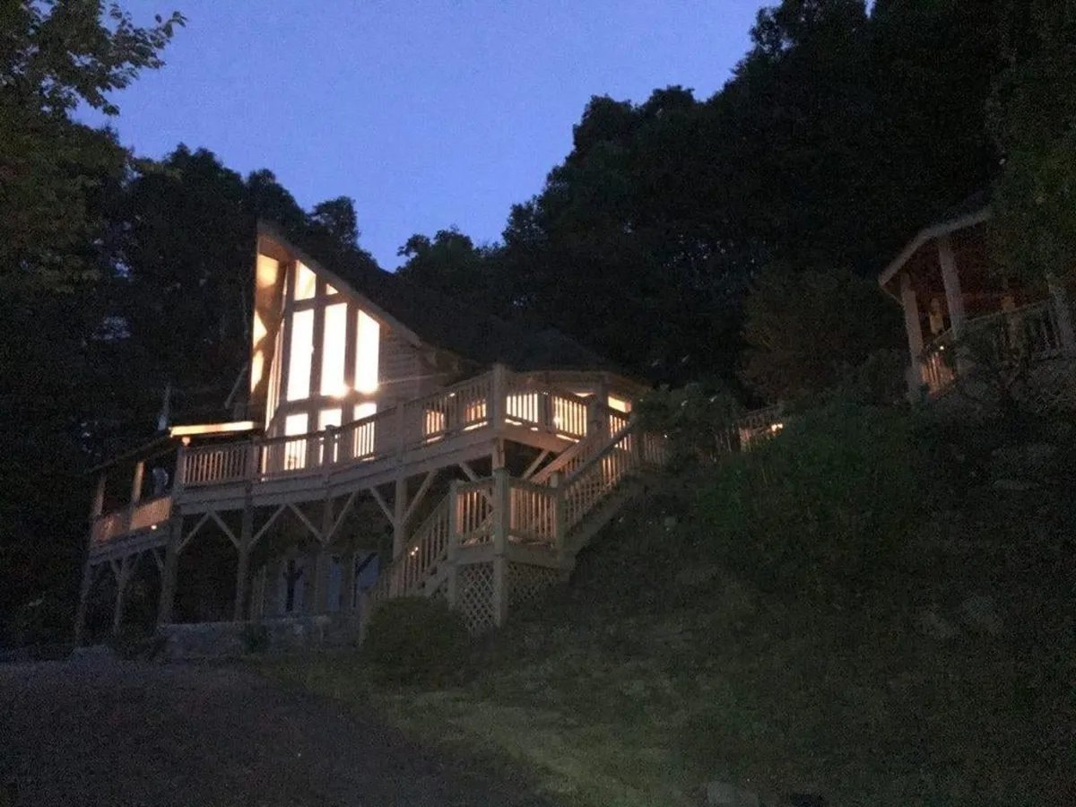 An exterior night-time view of the cabin, featuring its warmly illuminated structure, a spacious wooden deck with stairs, and surrounding trees. The shot captures the inviting glow of the cabin against the peaceful backdrop of the forest, emphasizing both the seclusion and the beauty of the natural setting.
