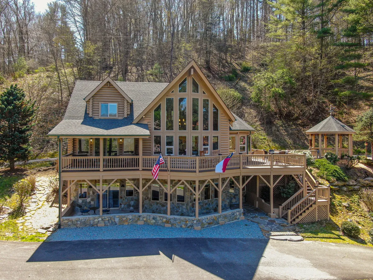 The image showcases the exterior of a beautifully renovated log cabin with a large front deck, expansive glass windows, sturdy stone foundation, and surrounded by lush Carolina trees. The setting radiates tranquility, and the deck is perfectly positioned for enjoying sunset views or morning coffee in nature.