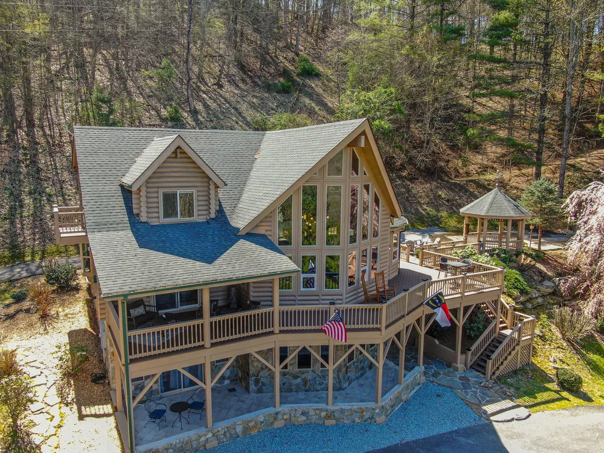The exterior of a charming log cabin with large windows and a welcoming deck area. Flags flutter by the stairs, and the expansive porch invites outdoor lounging, with greenery and mountains as a backdrop.