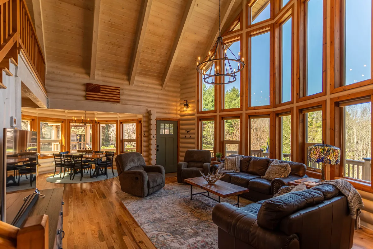 This image captures a wide view of the great room featuring expansive windows with mountain vistas, plush seating, exposed wooden beams, and the adjacent dining area. The inviting arrangement encourages family gatherings and conversation, with soft natural light filling the space.