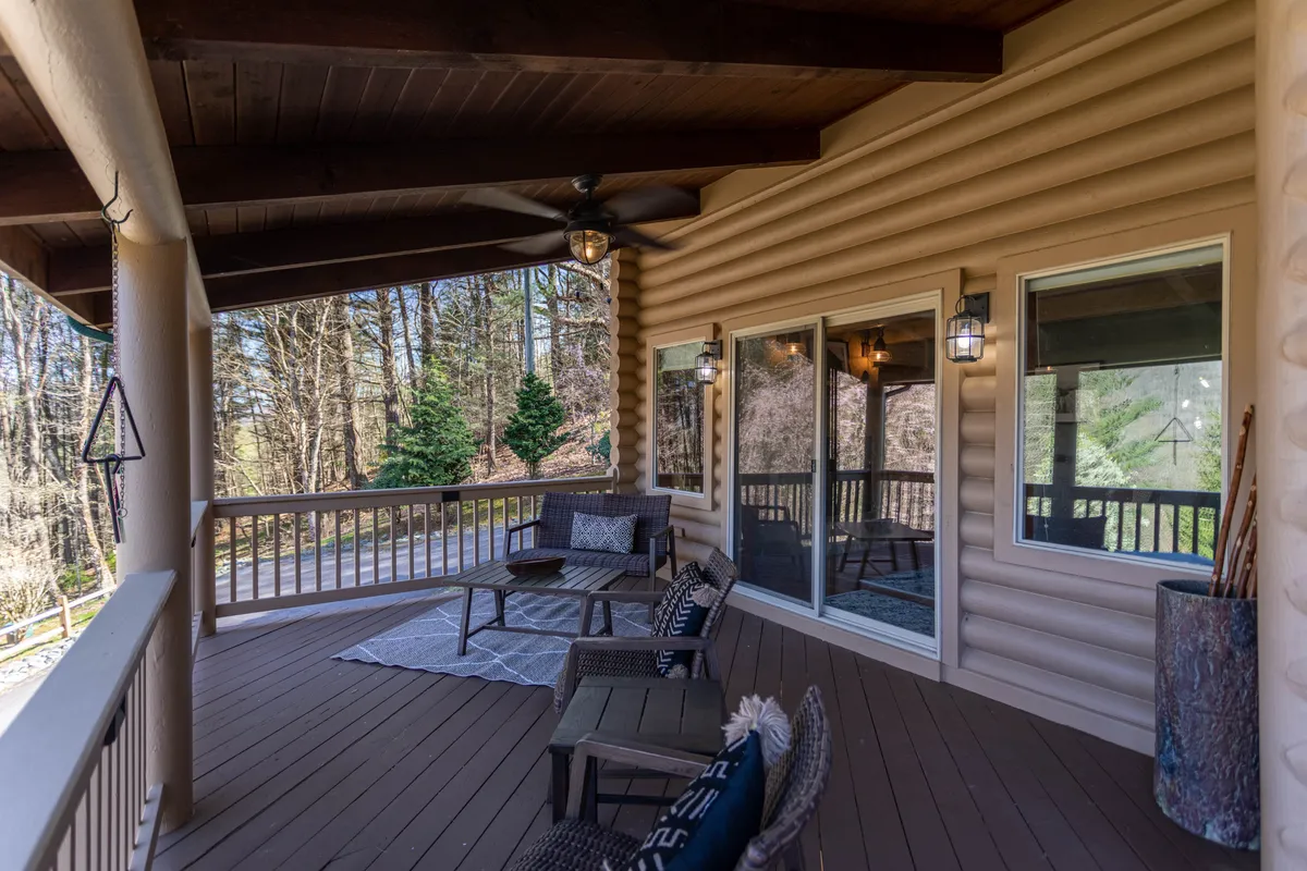 A covered, comfortable outdoor seating area features a wooden ceiling, decorative rug, and cozy furnishings, set against a backdrop of surrounding greenery. The space is designed for relaxation, reading, or gathering with friends, and is shaded yet open to nature.