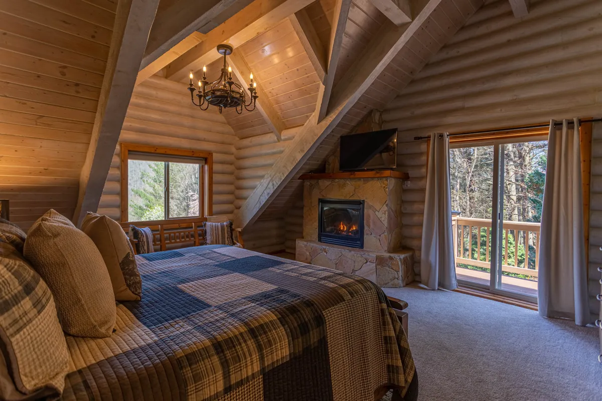 A spacious king suite with a king-sized bed adorned in plaid bedding, a stone fireplace, large window with scenic views, and a statement chandelier. The room exudes comfort and luxury, featuring both rustic accents and modern amenities like a TV.