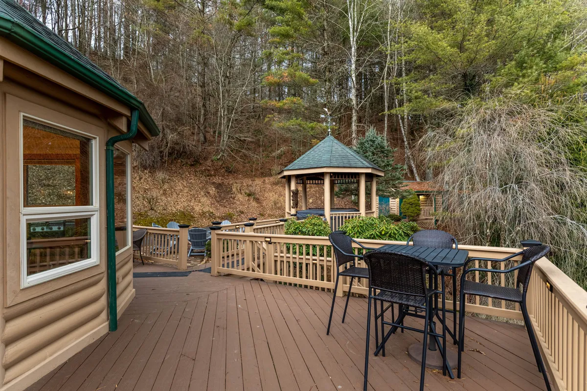 An inviting gazebo sits on a wooden deck surrounded by lush trees. The deck features comfortable seating, perfect for relaxing or socializing. The tranquil setting offers mountain air and privacy, with the gazebo likely housing the coveted hot tub mentioned in the section.